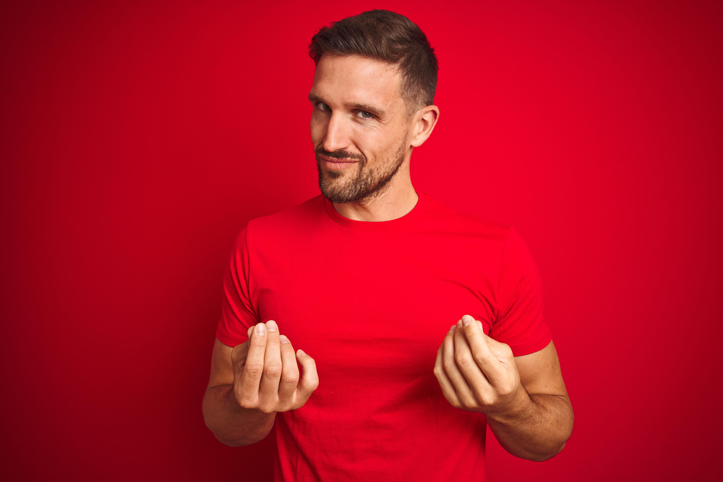Un homme en tee shirt rouge fait le signe "argent" avec ses doigts