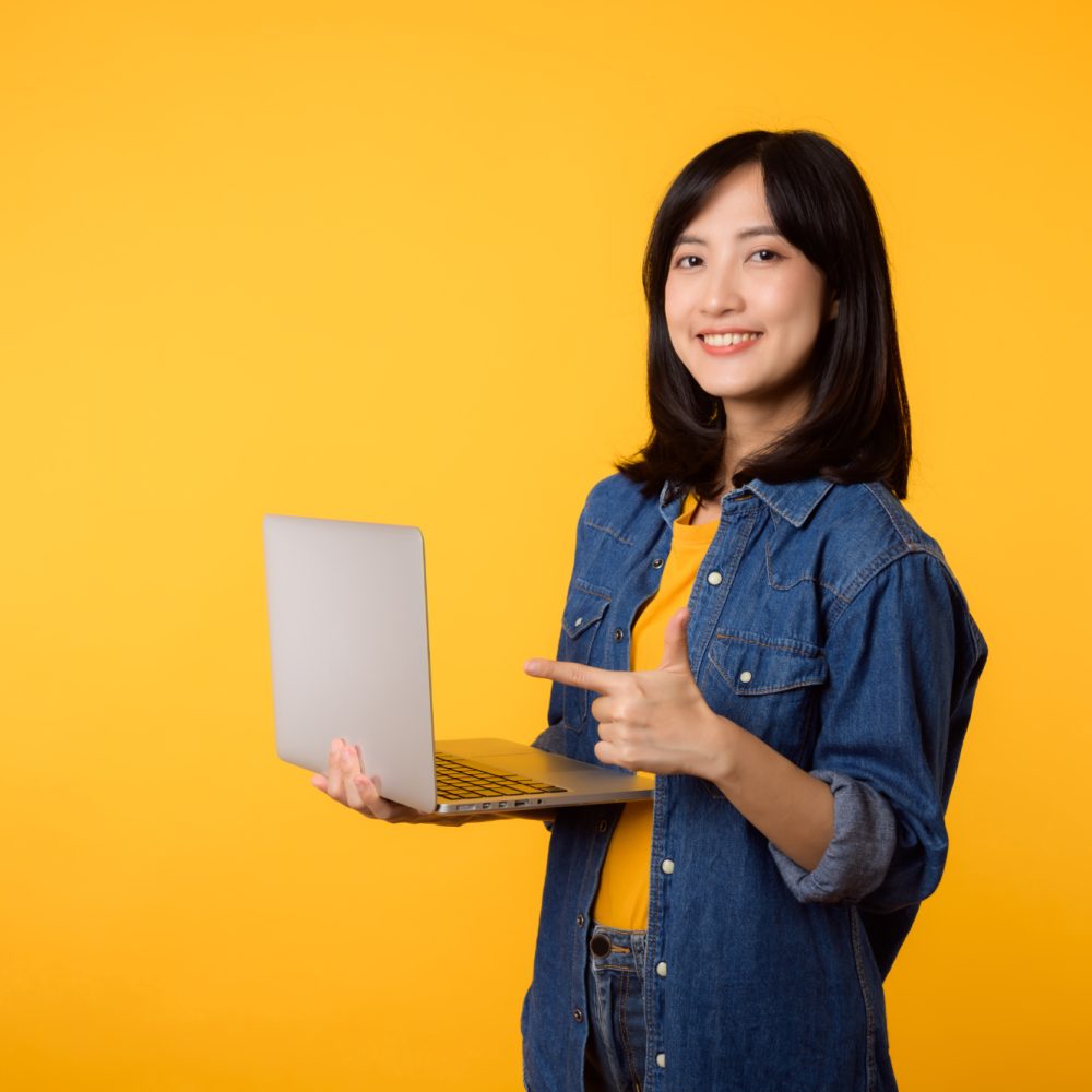 portrait happy young woman wearing yellow t-shirt and denim shirt holding laptop and point finger to screen isolated on yellow studio background. business technology application communication concept.